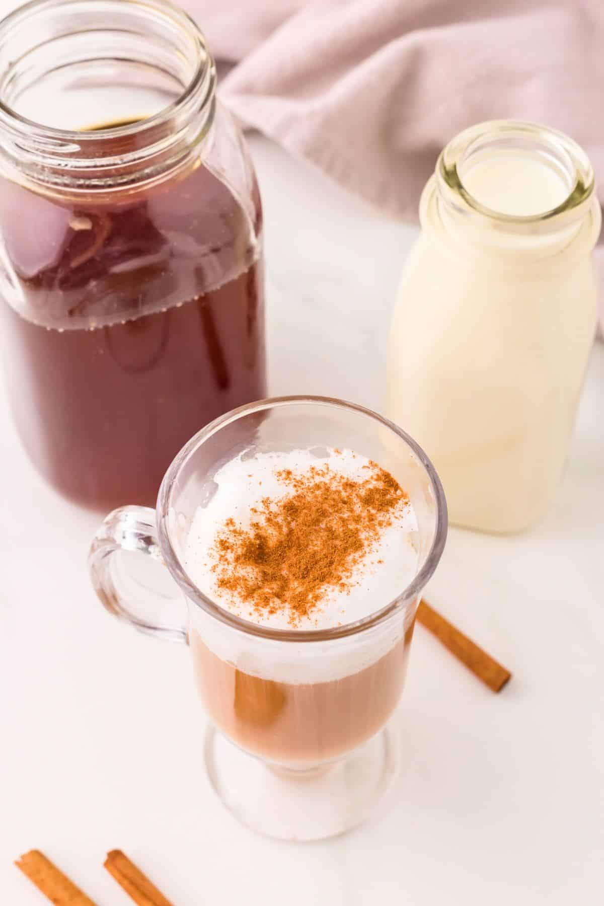 Chai tea latte in a glass mug topped with some ground cinnamon, with a bottle of chai concentrate and a bottle of milk behind. 