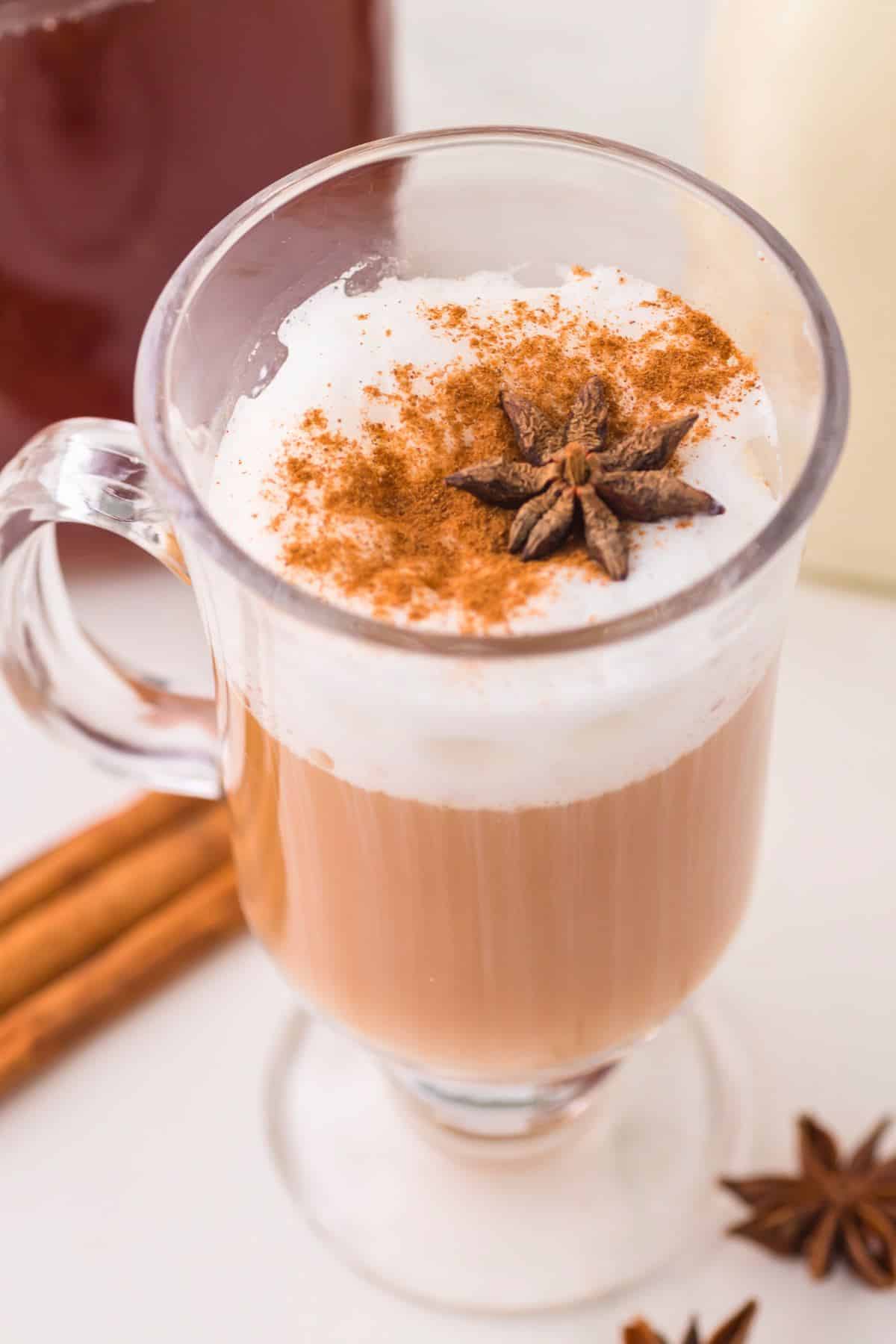Chai tea latte in a glass footed mug garnished with foamed milk a sprinkle of cinnamon and a star anise.