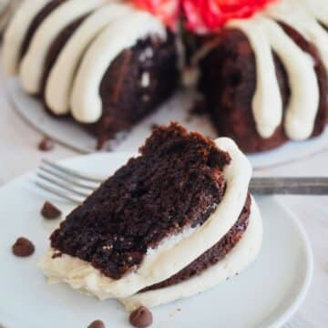 Rich slice of chocolate chocolate chip bundt cake on plate with fingers of frosting and full cake in background.