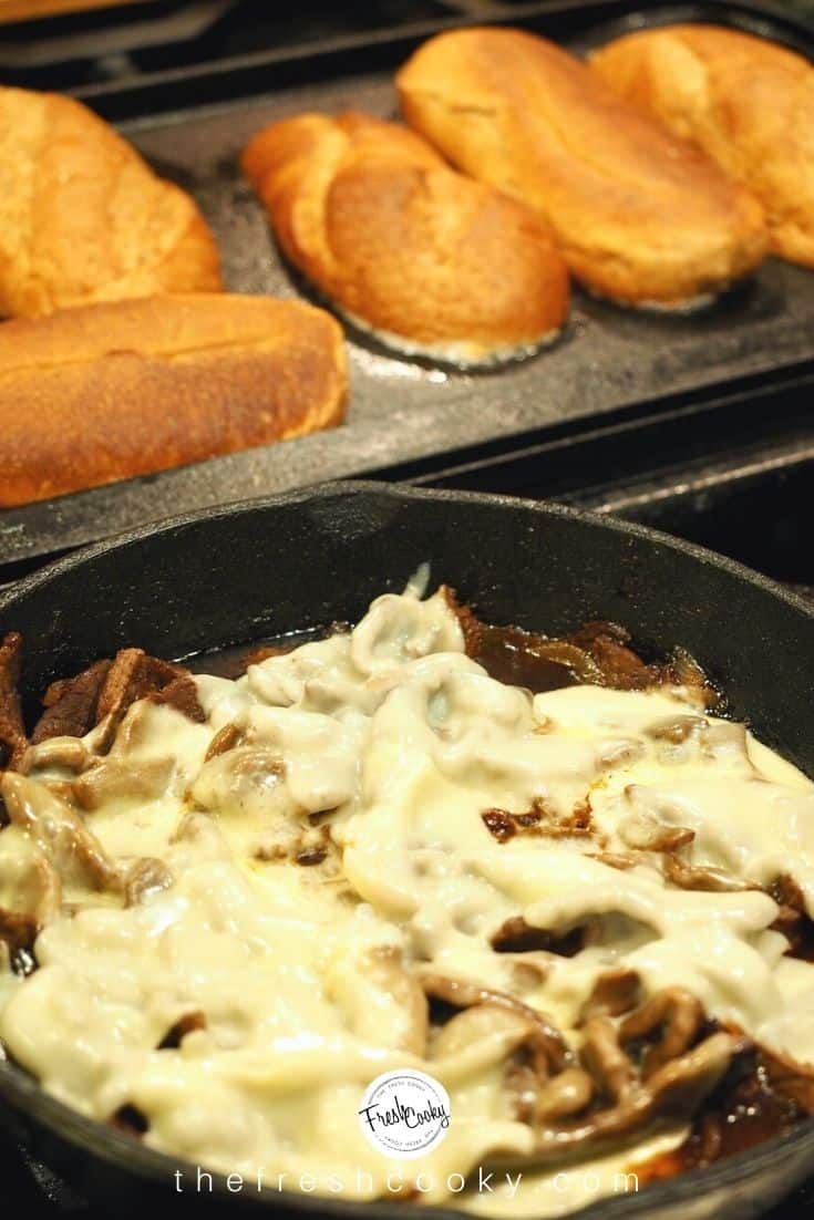 Skillet filled with melty cheesesteak with griddle in background toasting sub sandwich rolls.