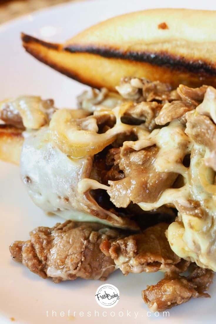 close up image of half cheesesteak sandwich on white plate, with toasted sub roll and stuffed with steak, melted cheese and sauteed onions. 