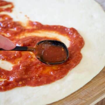 No rise pizza dough with hand spreading pizza sauce using a black spoon.
