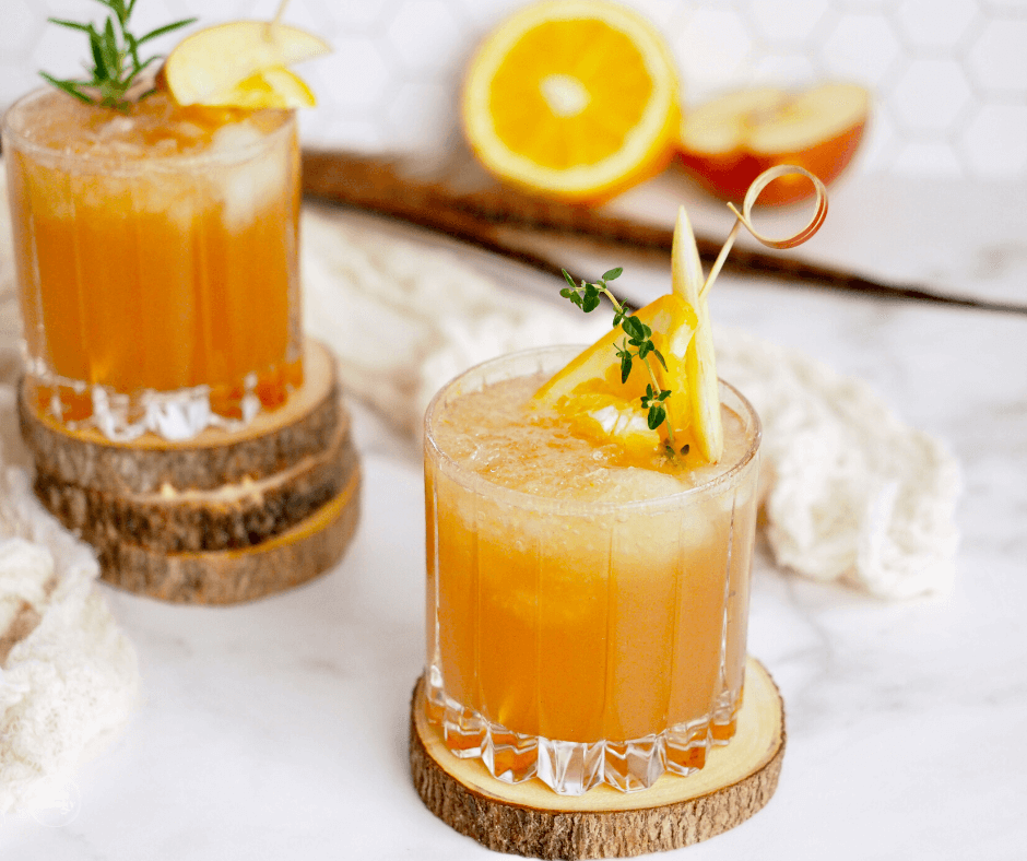 Apple bourbon cocktails in two glasses garnished with apples, herbs and sliced oranges.