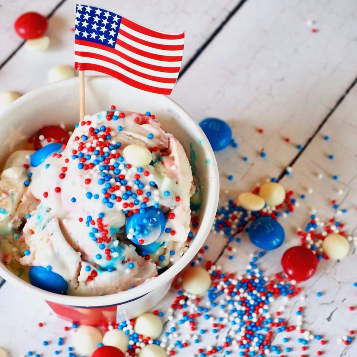 4th of July red white and blue ice cream in a flag cup with an American Flag pick, red, white and blue m & ms laying around with sprinkles.