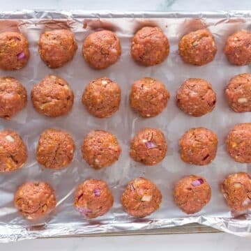 Raw ground beef with veggies is rolled into meatballs and laid on pan.