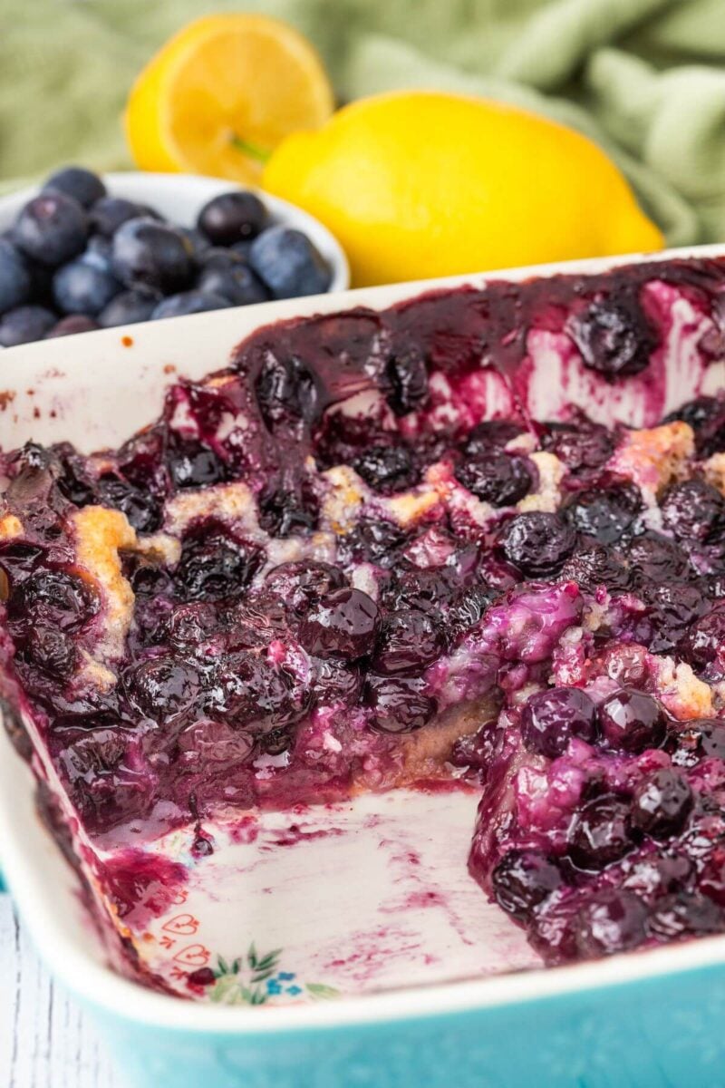 Melted baked blueberries show bright purple in the pan of cobbler next to fresh blueberries and lemons.