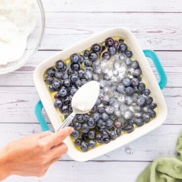 A hand uses a normal spoon to sprinkle sugar on top of a pan of blueberry dessert.