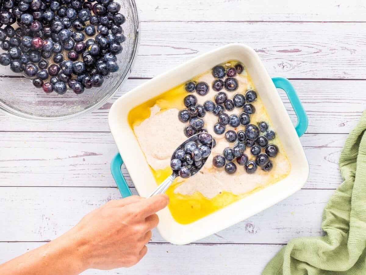 A hand uses a spoon to place blueberries on top of the unbaked cobbler ingredients in a square pan.