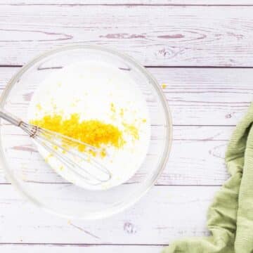 A whisk sits in a bowl of white sugar and bright yellow lemon zest.