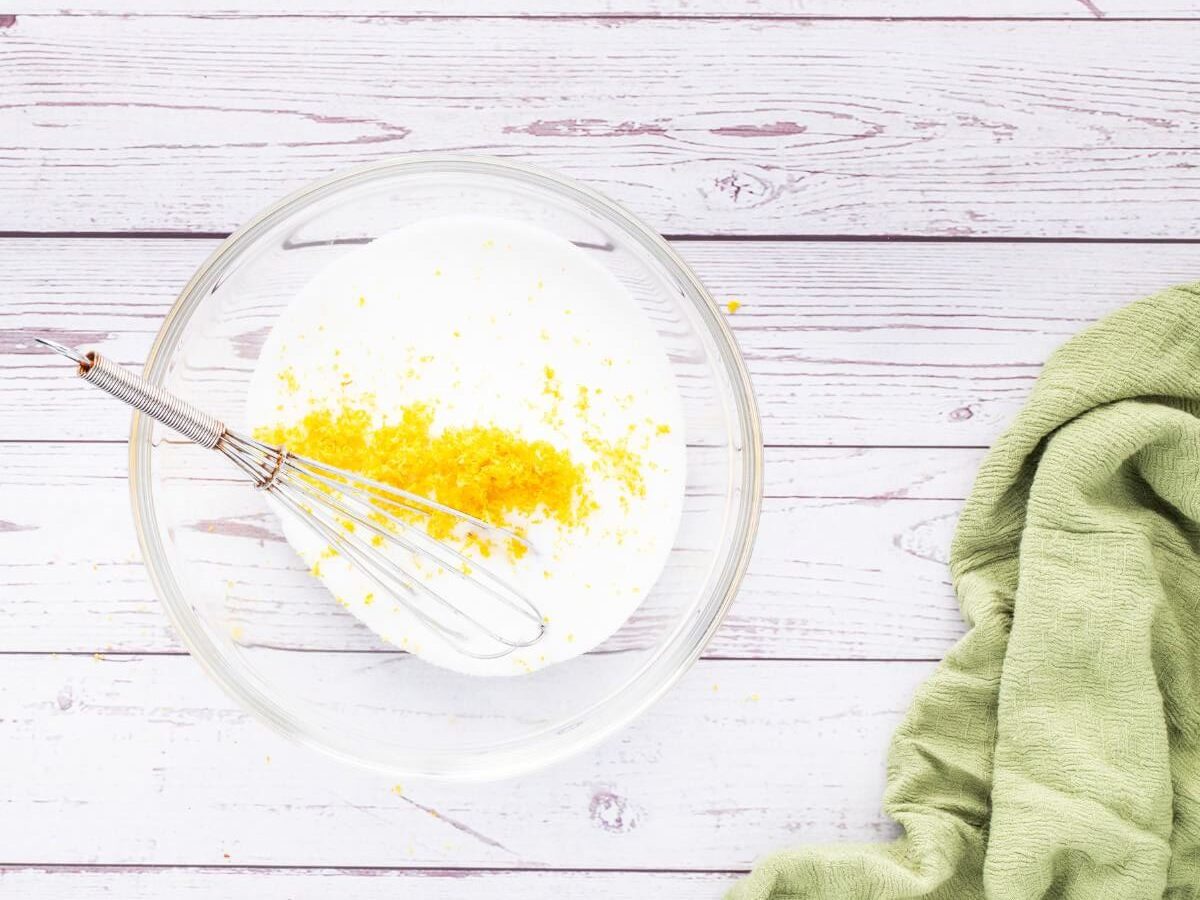 A whisk sits in a bowl of white sugar and bright yellow lemon zest.
