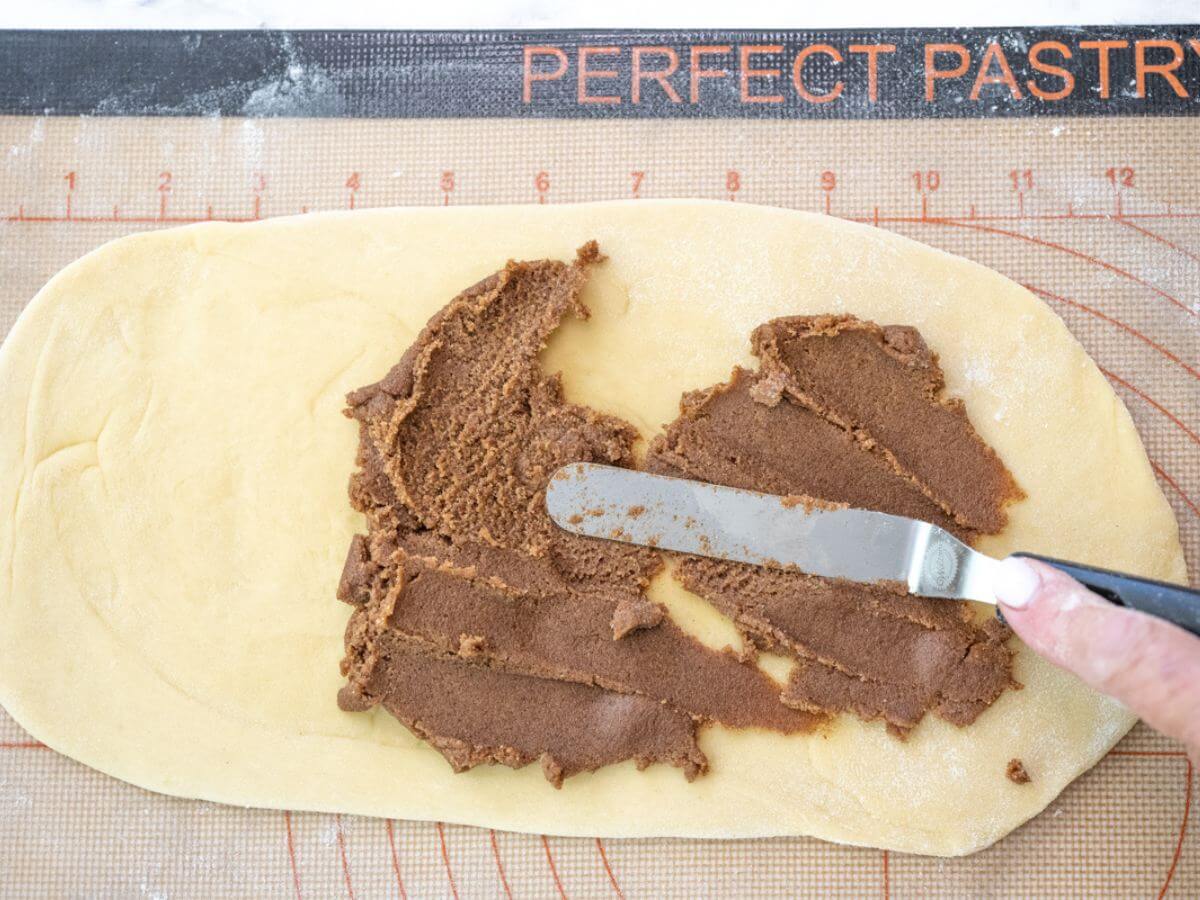 A hand is using a thin metal spatula to spread out the cinnamon filling on dough.