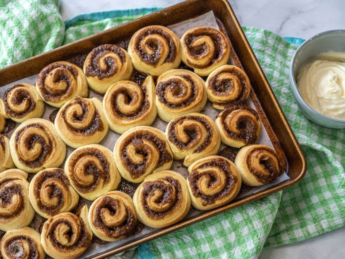 A baked pan of cinnamon buns sits on a checkered dishcloth by a bowl of icing.