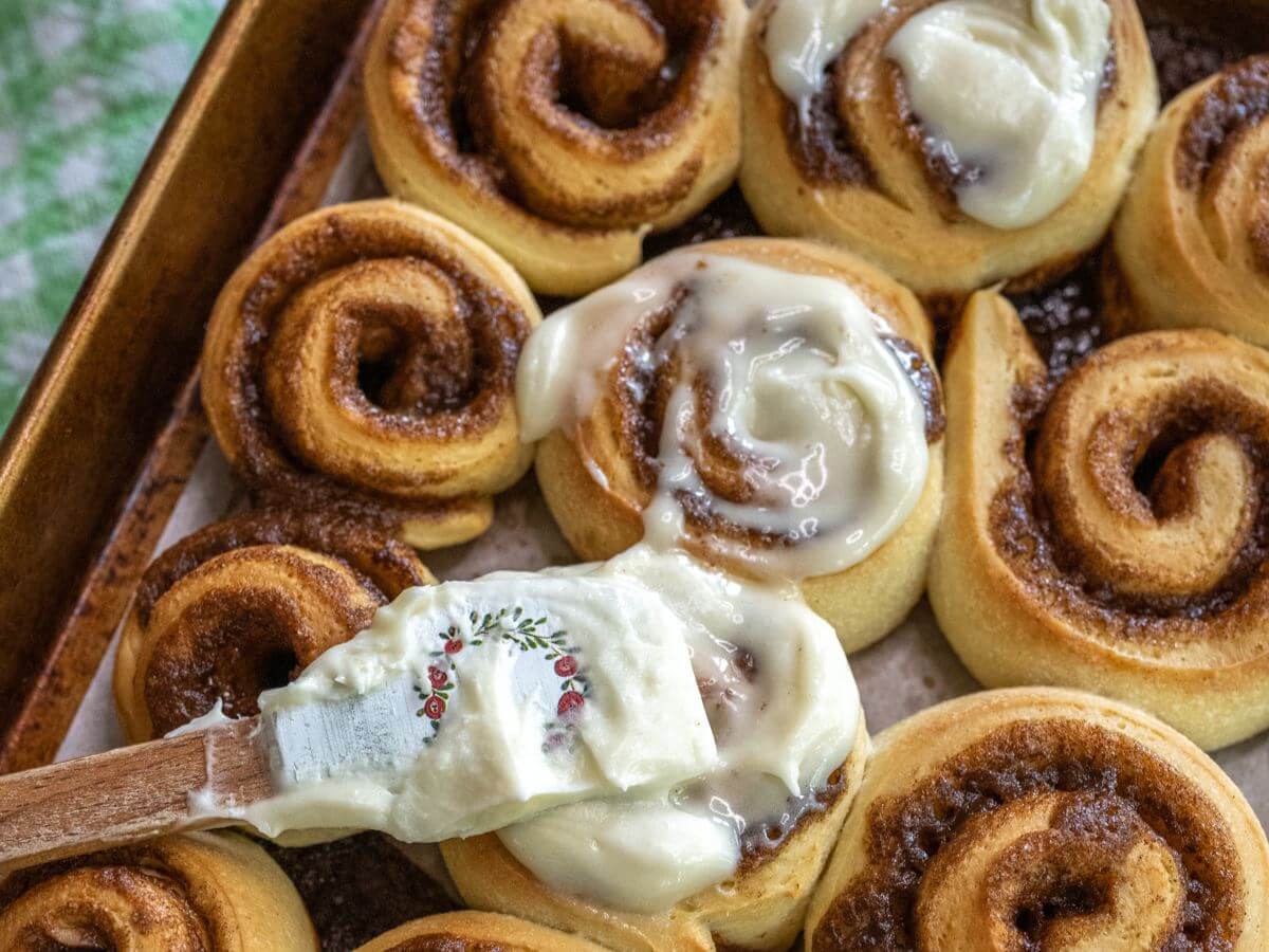 A close-up image of a spatula applying icing to baked cinnamon rolls shows a crunchy roll and smooth icing.