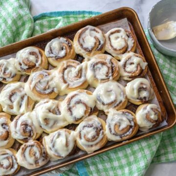An empty icing bowl and spatula sit next to a fully iced pan of tiny cinnamon buns.