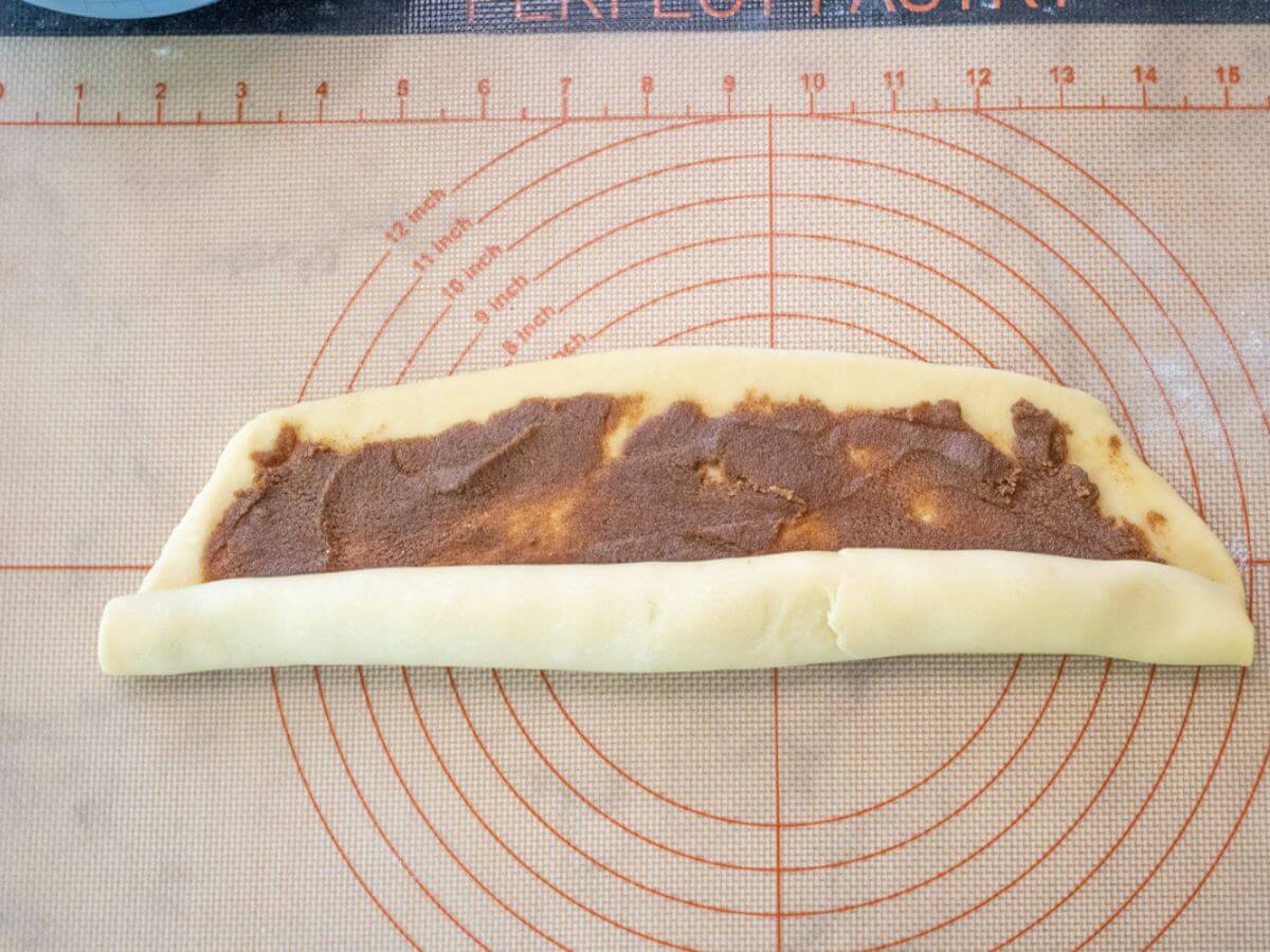Cinnamon filling is inside flat dough that is being rolled up.