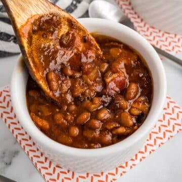 A wooden spoon placing cowboy baked beans into a serving bowl.