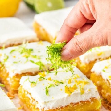 A hand sprinkles yellow and green zest from lemons and limes over white iced dessert bars.