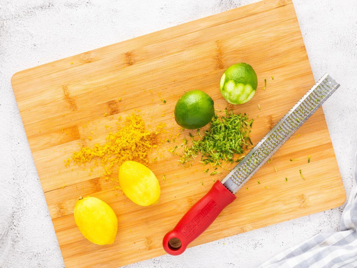 A zester lays next to partly zested lemons and limes on a wooden board.