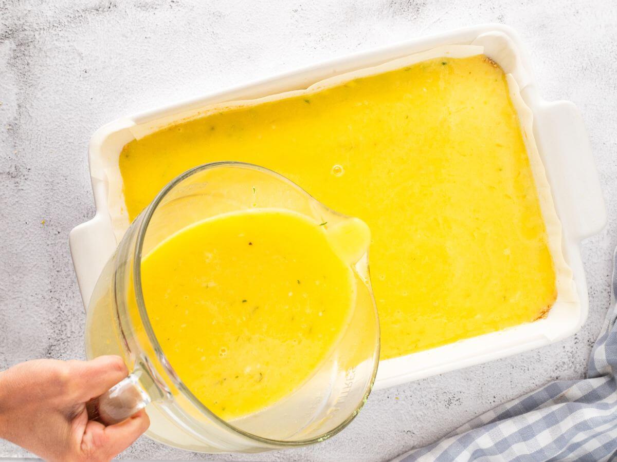 A hand pours a pitcher of yellow liquid filling mixture into baking pan.
