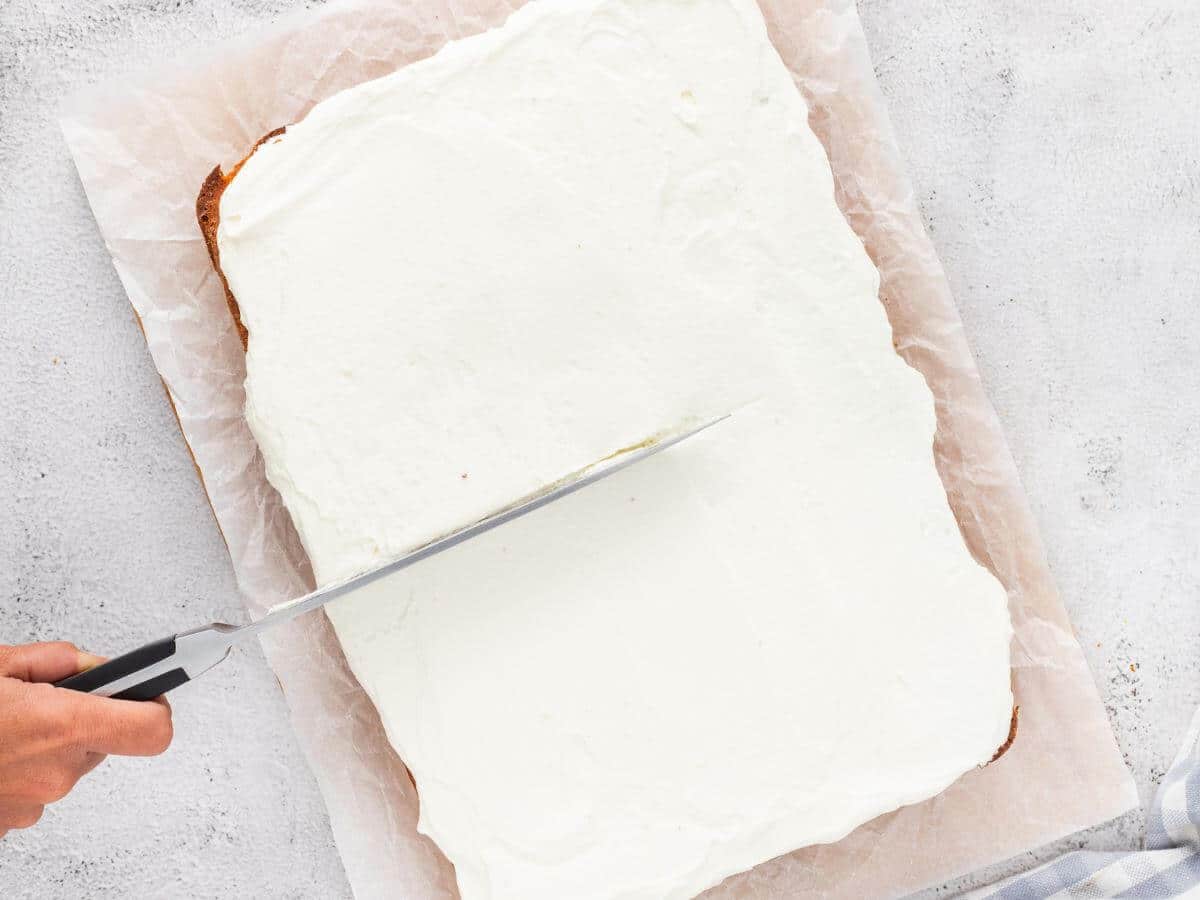 A white whip topped rectangle is cut down middle by a hand with a knife.