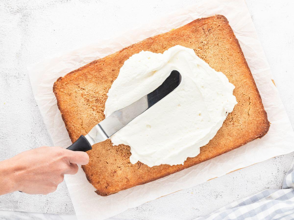 A hand uses a metal thin spatula to spread out whipped topping on fruit bars.