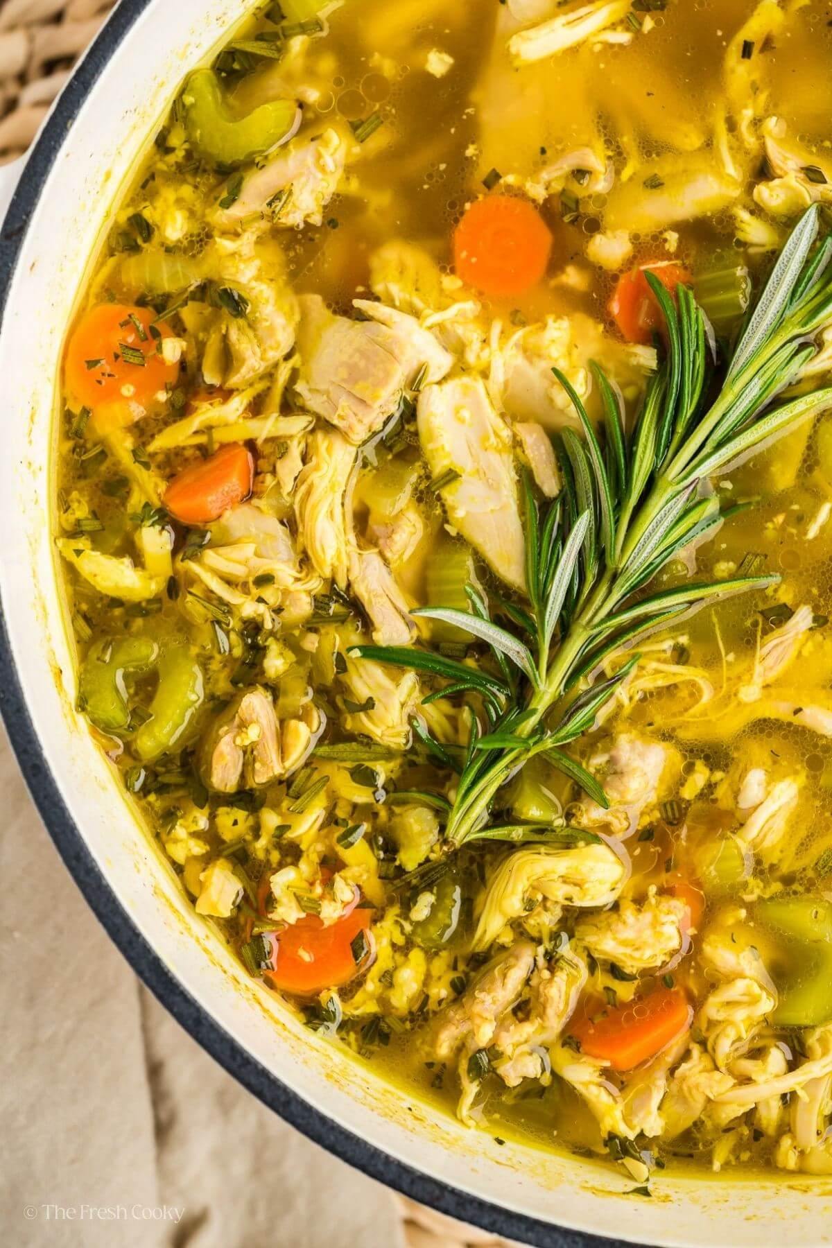 Close up shot of the chicken soup in a large pot topped with a sprig of rosemary.